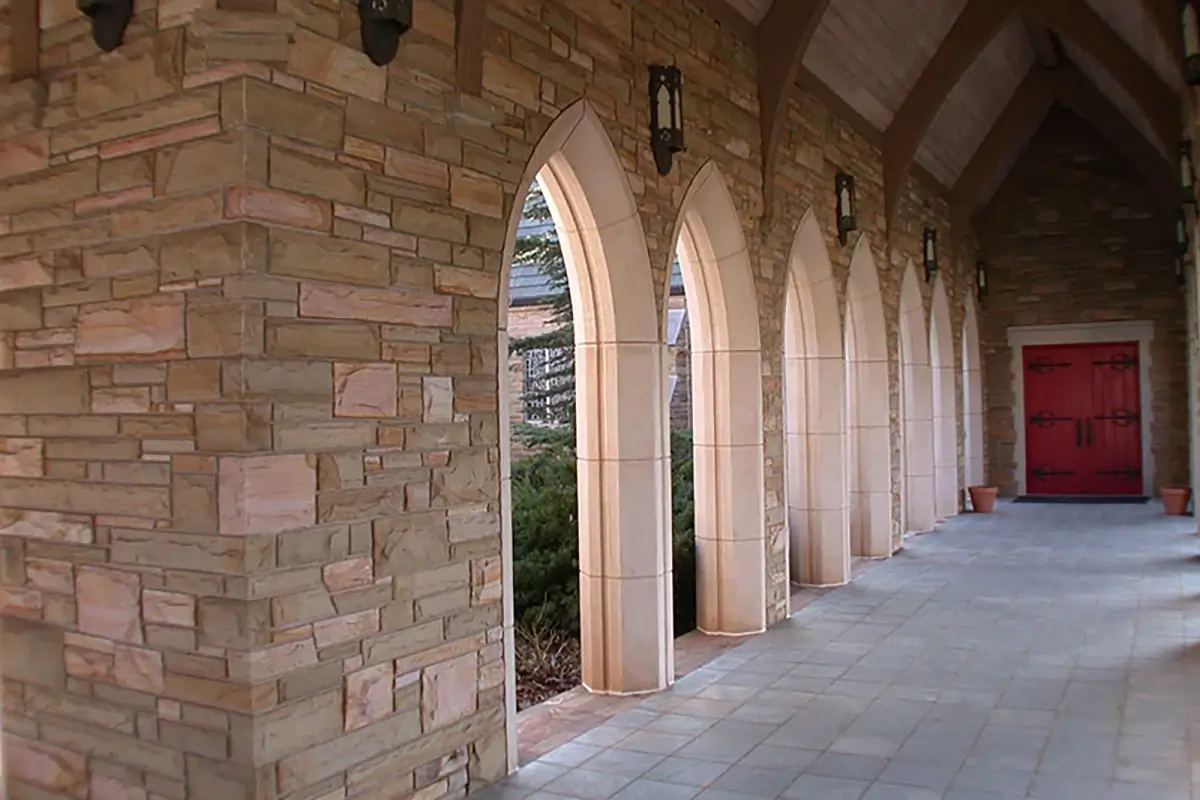 An outdoor stone cloister with a series of pointed arches leading to a bright red double door, featuring tan masonry walls, vaulted ceilings, and black lanterns.