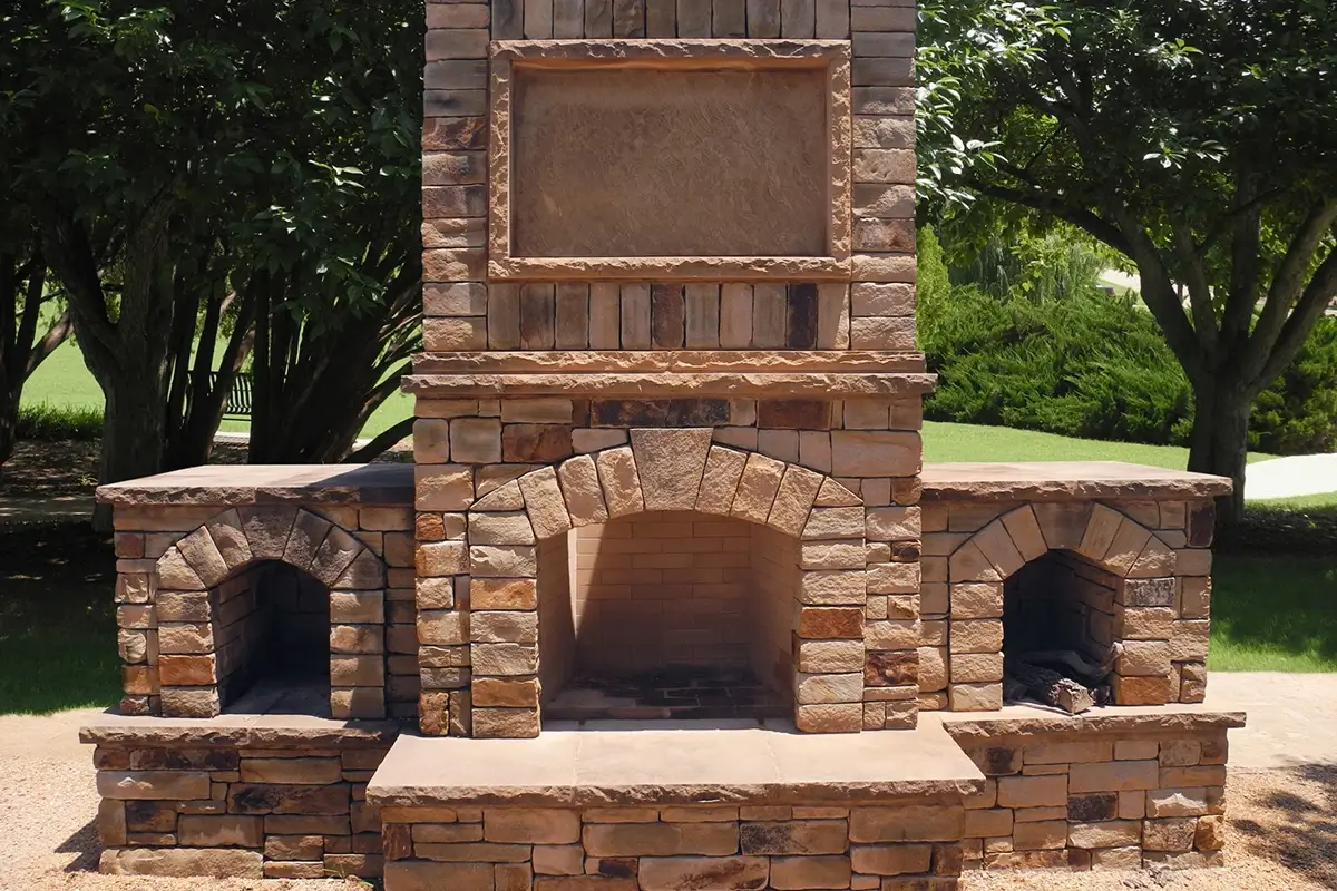 An outdoor stacked stone fireplace with a central arched hearth, two side wood storage nooks, and a tall chimney featuring a recessed rectangular stone panel.