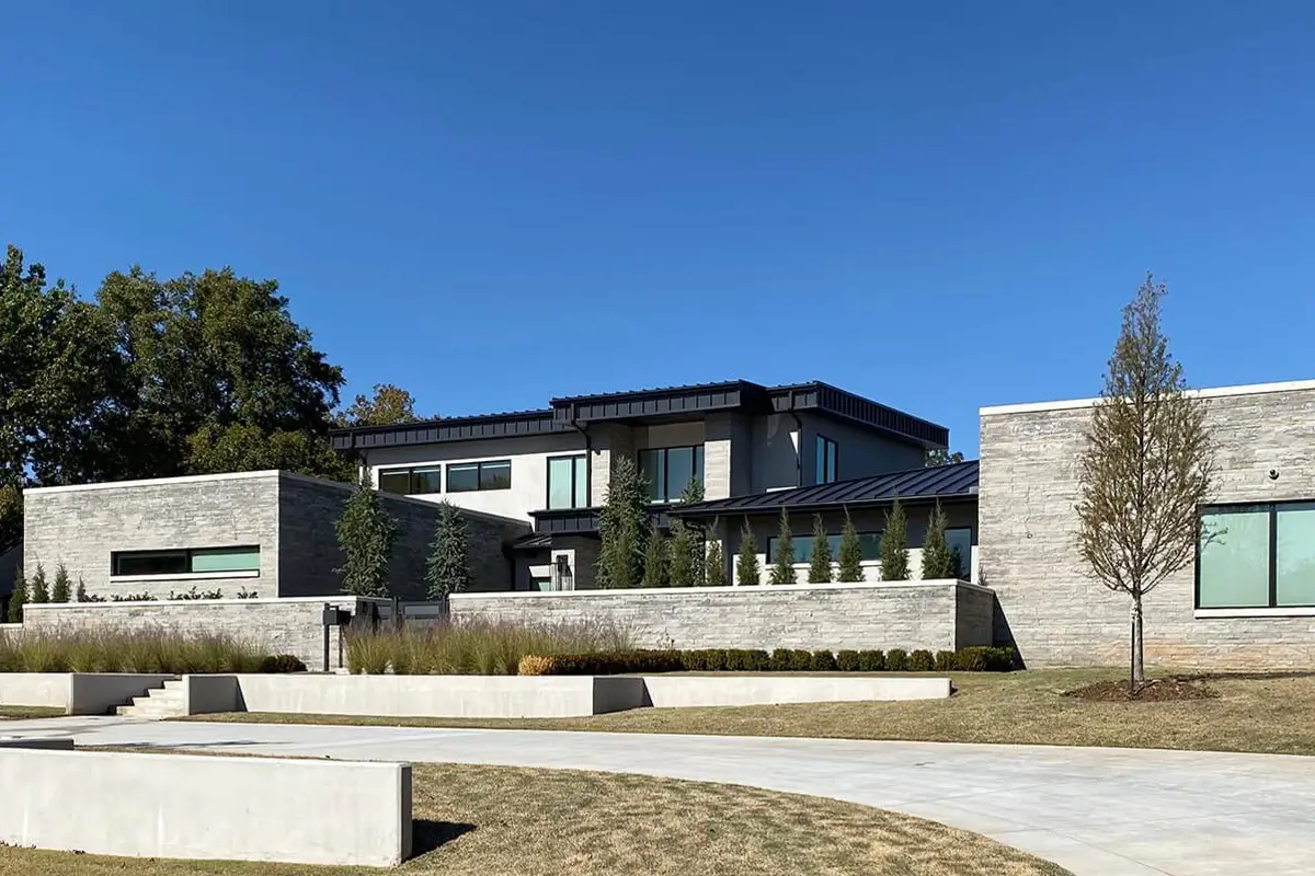 An expansive, modern two-story home featuring a combination of light gray stone and dark metal accents, set behind tiered concrete retaining walls and minimalist landscaping under a clear blue sky.