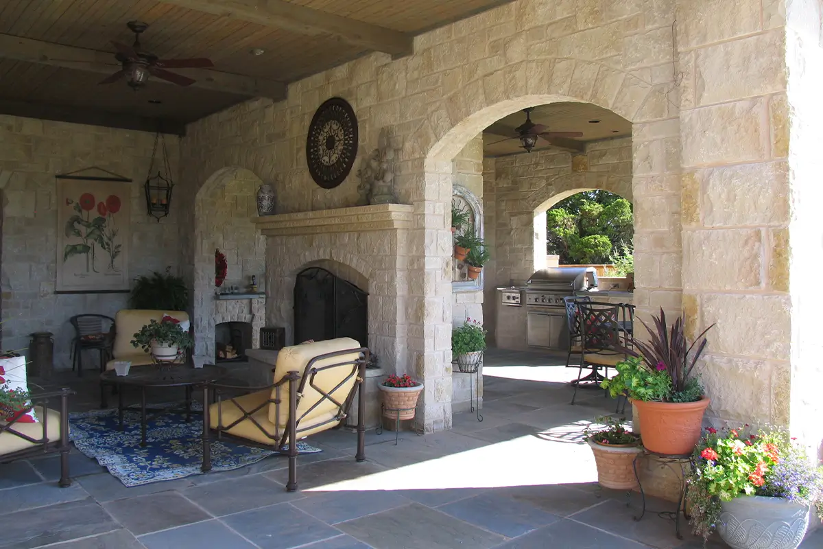 A luxurious covered stone patio featuring a large stone fireplace, yellow cushioned armchairs on a patterned rug, and a stone archway leading to an outdoor kitchen grill station.