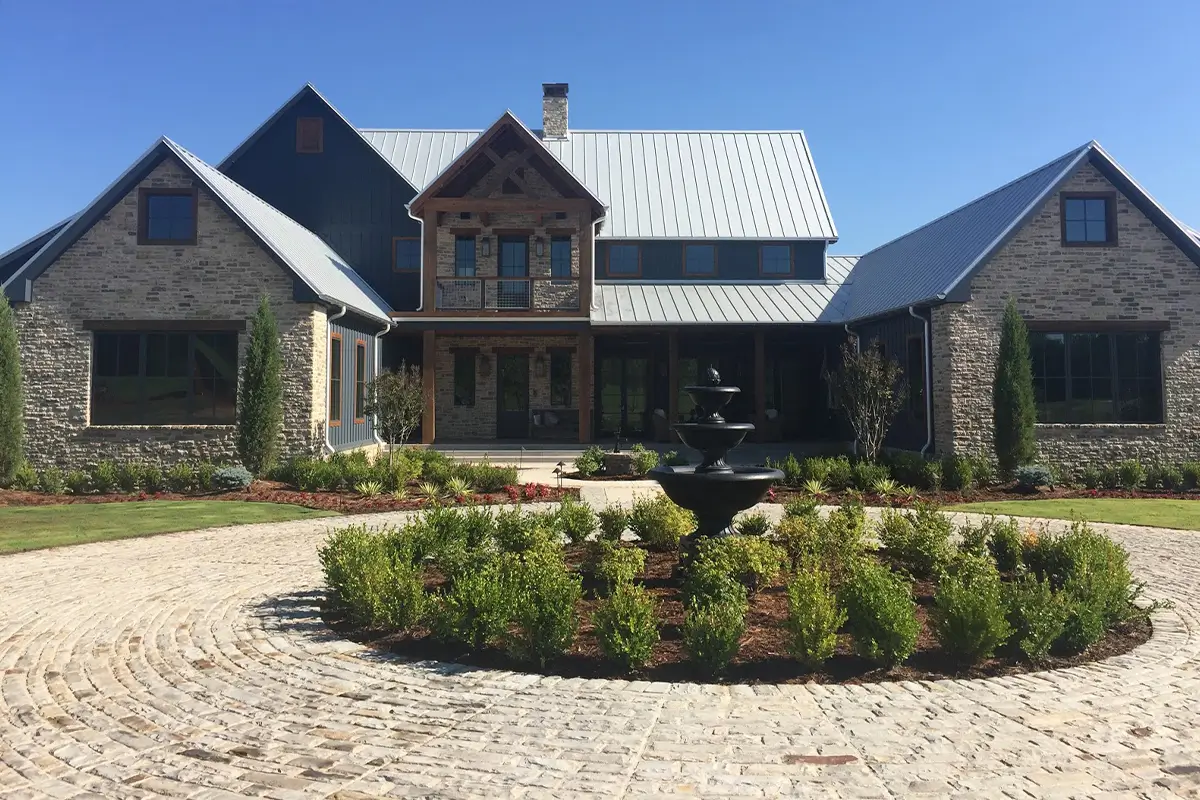 A large modern stone estate with a circular cobblestone driveway and a central fountain under a clear blue sky.